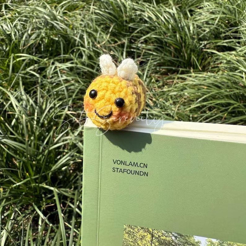 Puede incluir: Un marcap&aacute;ginas de abeja de ganchillo, amarillo y marr&oacute;n, con una cara sonriente, ojos negros y antenas blancas. La abeja descansa sobre un libro verde. El texto "VONLAM.CN STAFOUNDN" es visible.