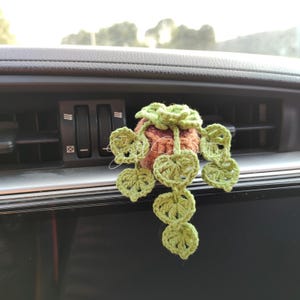 以下が含まれることがあります： 手作りの、小さな植物の形をしたかぎ針編みの車の芳香剤。茶色のポット、緑の葉、緑の花があります。芳香剤は車の通気口から吊り下げられています。