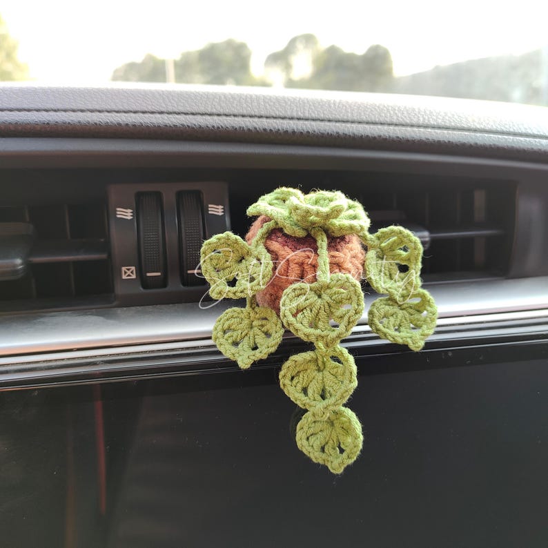 以下が含まれることがあります： 手作りの、小さな植物の形をしたかぎ針編みの車の芳香剤。茶色のポット、緑の葉、緑の花があります。芳香剤は車の通気口から吊り下げられています。
