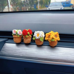 May include: Four crochet flower pot decorations. Each pot is a different flower: pink and white roses, red roses, a white daisy, and yellow sunflowers. The pots are brown and sit on a silver surface.