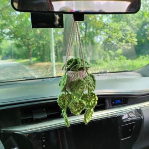 Könnte beinhalten: Gehäkelte Hängepflanze zur Dekoration für das Auto. Der Topf ist hellbeige, die Blätter grün. Aufgehängt an einer beigen Schnur, verleiht sie dem Innenraum des Autos einen Hauch von Natur. Ein charmantes Accessoire.