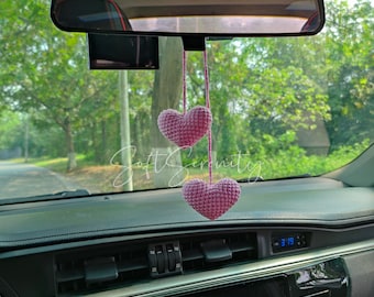 May include: A car interior with a rearview mirror and dashboard. Two pink crocheted hearts hang from the mirror. The text "Soft Serenity" is written across the image.
