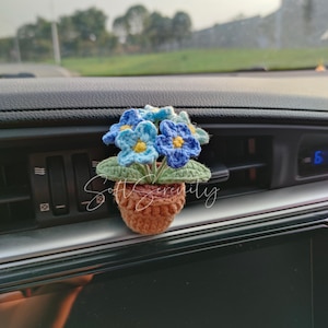 以下が含まれることがあります： 青と黄色の花と緑の葉が付いた、茶色のポットに入ったかぎ針編みのフラワーアレンジメント。「Soft Serenity」の文字が前面に書かれています。アレンジメントは車のダッシュボードに配置されています。