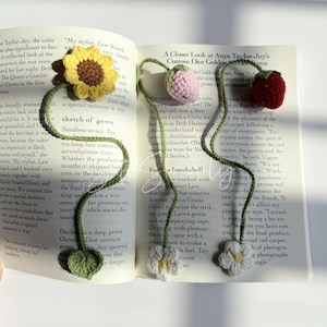 Puede incluir: Marcadores florales de ganchillo en un libro. Los marcadores presentan un girasol, un tulip&aacute;n rosa, una fresa roja y dos flores blancas con tallos y hojas verdes. El libro est&aacute; abierto en una p&aacute;gina con texto.