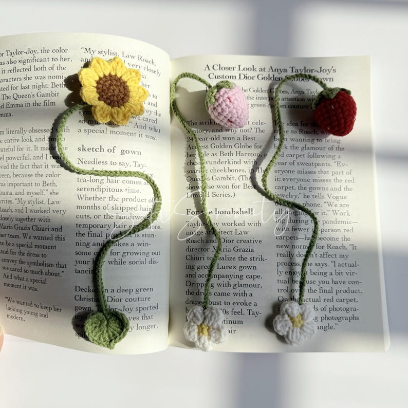 Puede incluir: Marcadores florales de ganchillo en un libro. Los marcadores presentan un girasol, un tulip&aacute;n rosa, una fresa roja y dos flores blancas con tallos y hojas verdes. El libro est&aacute; abierto en una p&aacute;gina con texto.