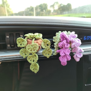 以下が含まれることがあります： 手作りのかぎ針編みの車の通気口の装飾。1つはハート型の葉と茶色のポットが付いた緑色のつるです。もう1つは紫と緑の花のクラスターです。手作りの装飾。