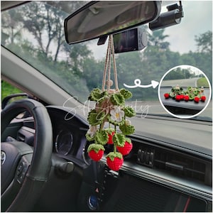 Può includere: Un ciondolo per auto all'uncinetto fatto a mano con un design a pianta di fragola. Il ciondolo include fragole rosse, foglie verdi e fiori bianchi, appesi a una corda beige. Il ciondolo è esposto all'interno di un'auto.