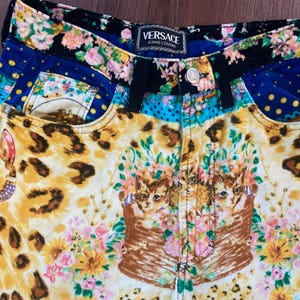 Peut inclure: Pantalon en jean Versace Jeans Couture au design vibrant et &eacute;clectique. Le pantalon pr&eacute;sente un m&eacute;lange de motifs, notamment un imprim&eacute; l&eacute;opard, des compositions florales et un motif de chat. La taille porte une &eacute;tiquette Versace.