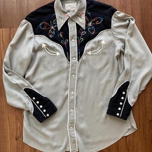 Pode incluir: Camisa vintage em estilo Western cinza com detalhes em preto. A camisa apresenta desenhos florais bordados coloridos no peito e nos punhos. Tubulação branca delineia a gola, a frente e os punhos. Os punhos são pretos com botões brancos.