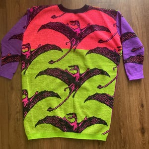 May include: A long-sleeved sweater with a vibrant design. The sweater features a color-blocked pattern with neon pink and green sections. Black pterodactyls with pink accents are scattered across the front. The sleeves are purple. The sweater is made of a knitted material.