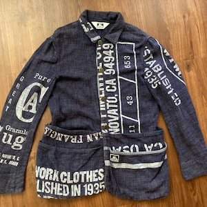 May include: A navy blue work jacket with white printed text and graphics. The jacket features long sleeves, two front pockets, and a button-up front. Text includes "WORK CLOTHES ESTABLISHED IN 1935" and "NOVATO, CA 94949".