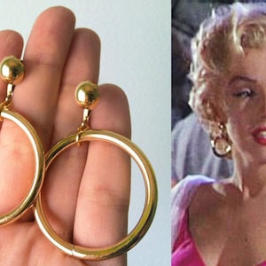 May include: A pair of gold clip-on earrings with large round hoops. The earrings are shown on a hand and in a close-up of Marilyn Monroe wearing them.