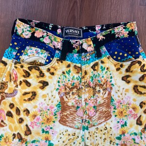 Peut inclure: Short en jean Versace Jeans Couture au design vibrant et &eacute;clectique. Le short pr&eacute;sente un m&eacute;lange de motifs, notamment des imprim&eacute;s l&eacute;opard, des compositions florales et des illustrations de chats. La ceinture et les poches ont des motifs floraux et &agrave; pois contrast&eacute;s.