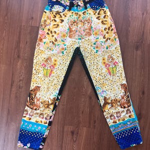 Peut inclure: Jean taille haute &agrave; motifs avec un design unique. Le jean pr&eacute;sente un fond jaune avec un imprim&eacute; l&eacute;opard, des motifs floraux et des illustrations de chats. La ceinture pr&eacute;sente un motif floral et bleu, avec le logo Versace visible.