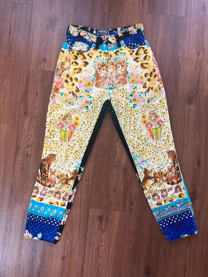 Peut inclure: Jean taille haute &agrave; motifs avec un design unique. Le jean pr&eacute;sente un fond jaune avec un imprim&eacute; l&eacute;opard, des motifs floraux et des illustrations de chats. La ceinture pr&eacute;sente un motif floral et bleu, avec le logo Versace visible.