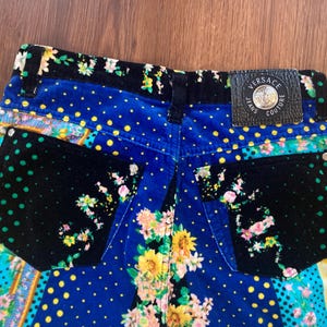 Peut inclure: Gros plan sur l'arri&egrave;re d'un jean Versace color&eacute;. Le jean pr&eacute;sente un motif patchwork avec des motifs floraux, &agrave; pois et animaliers. Un patch en cuir noir avec le logo Versace Jeans Couture est visible.
