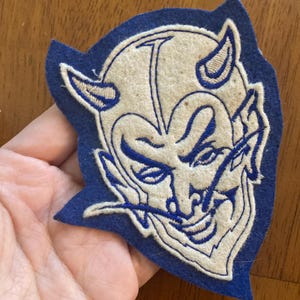May include: A blue and white felt patch featuring a devil mascot design. The devil has a stern expression with horns and a pointed chin. The patch is a vintage style, likely for a sports team or school.