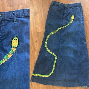 Puede incluir: Falda larga de mezclilla azul oscuro con un diseño de serpiente bordada. La serpiente es amarilla, verde y negra. La falda tiene cintura alta y una silueta acampanada, con cierre de botón y cremallera.