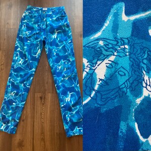 May include: High-waisted blue jeans with a vibrant, abstract water-like pattern in shades of blue and white. The jeans feature a classic five-pocket design and a button closure. A close-up reveals a Medusa head design.