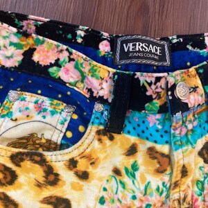 Peut inclure: Gros plan sur un short en jean Versace Jeans Couture color&eacute;. Le short pr&eacute;sente un motif patchwork avec des motifs floraux, l&eacute;opard et &agrave; pois. La ceinture et les poches sont orn&eacute;es de boutons argent&eacute;s. L'&eacute;tiquette Versace Jeans Couture est visible.