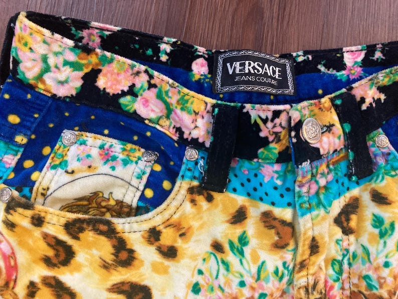 Peut inclure: Gros plan sur un short en jean Versace Jeans Couture color&eacute;. Le short pr&eacute;sente un motif patchwork avec des motifs floraux, l&eacute;opard et &agrave; pois. La ceinture et les poches sont orn&eacute;es de boutons argent&eacute;s. L'&eacute;tiquette Versace Jeans Couture est visible.