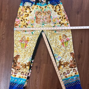 Peut inclure: Jean taille haute avec un motif vibrant. Le jean pr&eacute;sente un m&eacute;lange d'imprim&eacute; l&eacute;opard, de motifs floraux et de chats. La ceinture porte le logo "Versace". La palette de couleurs comprend le jaune, le bleu, le noir et le rose.