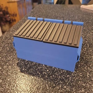 Puede incluir: Una caja de almacenamiento plegable azul con una parte superior negra acanalada. La caja está hecha de plástico y tiene un diseño simple.