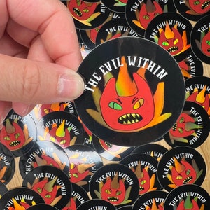 May include: A pile of round stickers with a black background and a red, yellow, and orange cartoon monster with the text "The Evil Within" in white.