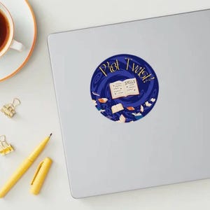 May include: A sticker with a blue and gold design featuring the words "Plot Twist?" and "Spells" around an open book. The sticker is on a laptop next to a cup of coffee, a yellow pen, and gold binder clips.
