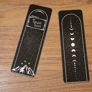 May include: Two black bookmarks with white designs. One features the text "Welcome to The Night Court" above a mountain scene and stars. The other has a moon phase design. Both have a starry background.