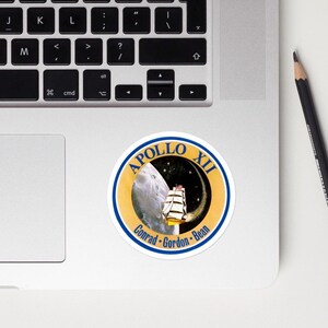 May include: A round sticker featuring the Apollo XII mission logo. The design includes a depiction of the moon, a sailing ship, and the names Conrad, Gordon, and Bean. The sticker has a blue and gold color scheme.
