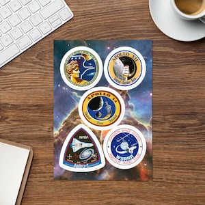 May include: A collection of space-themed stickers featuring NASA logos and mission patches. The stickers include the Apollo missions, with detailed illustrations and text. The background is a colorful nebula, and the stickers are arranged on a wooden surface.