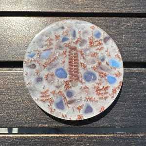 May include: A round, white resin art piece with embedded blue and purple stones, copper wire accents, and a central copper coil. The piece is set on a dark wooden surface, showcasing its intricate details and textures.