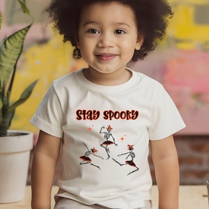 May include: A white t-shirt with the phrase "Stay Spooky" in orange and black lettering. The shirt features a graphic of three dancing skeletons wearing tutus and witch hats. The child is smiling.
