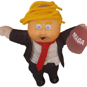 May include: A Cabbage Patch Kid doll with a yellow wig, wearing a black suit and red tie, holding a red hat with the word "MAGA" printed on it.