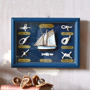May include: A framed nautical display featuring a central model sailboat surrounded by eight white rope knots on a dark blue background. The frame is blue, and each knot is labelled with a small gold plaque. The display is mounted on a wall.