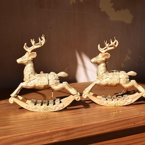 May include: Two gold-toned rocking deer figurines, each featuring a detailed saddle and antlers. The deer are mounted on curved rocking bases decorated with leaf motifs and small metallic bars, creating a cohesive pair of decorative ornaments.