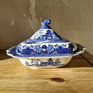Puede incluir: Una fuente de cerámica azul y blanca con tapa. El plato presenta un diseño de paisaje detallado con pagodas y árboles. La tapa tiene un asa decorativa. El plato está colocado sobre una superficie de madera.
