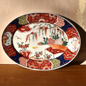 May include: Oval ceramic serving platter with a detailed Japanese-style design. The rim is dark blue with red and white floral patterns. The center features a landscape with flowers, birds, and a red structure.