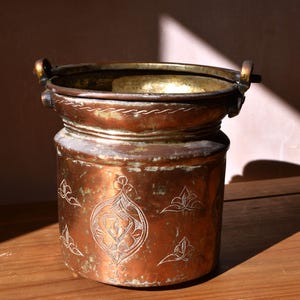 May include: A cylindrical copper bucket featuring a wide rim with two side handles. The exterior surface displays a weathered patina and is decorated with etched floral motifs and a central medallion design, showcasing a warm metallic finish.