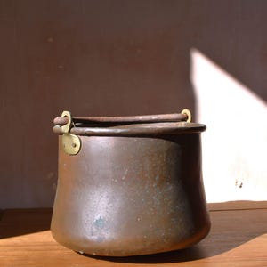 May include: A round copper pot with a wide opening and a slightly curved body. The pot features a dark, weathered patina with subtle green oxidation spots. It includes a thick, dark metal swing handle attached by two brass-coloured metal brackets.