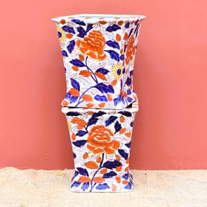 Puede incluir: Dos jarrones de cerámica cuadrados apilados. Los jarrones son blancos con un estampado floral con flores naranjas, hojas azules y detalles amarillos. Los jarrones tienen una parte superior ligeramente ensanchada y una base cuadrada.