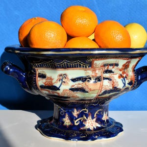 May include: A blue and white ceramic fruit bowl filled with oranges and a lemon. The bowl has a pedestal base and handles. The bowl is decorated with a landscape scene in red, white, and blue.