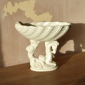 May include: Cream-coloured ceramic centerpiece bowl with a shell-like design. The bowl is supported by a base featuring sculpted figures. The bowl has a textured, leaf-like pattern on the exterior. The piece is displayed on a wooden surface.