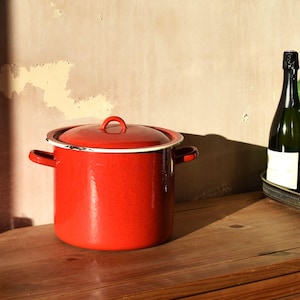 May include: A bright red enamel cooking pot with a lid and white trim. The pot has two side handles and a loop handle on the lid. It is on a wooden surface, with a bottle of wine in the background.