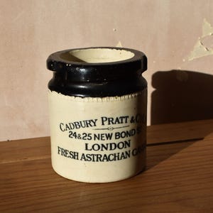 Può includere: Un vaso vintage color crema con bordo nero, con la scritta "CADBURY PRATT & Co. 24 & 25 NEW BOND ST LONDON FRESH ASTRACHAN CIDER" in nero. Il vaso cilindrico è appoggiato su una superficie di legno.