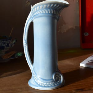 Puede incluir: Jarra de cerámica azul claro con cuerpo cilíndrico y asa curva. La jarra presenta motivos decorativos de hojas en la parte superior e inferior. Se encuentra sobre una superficie de madera.