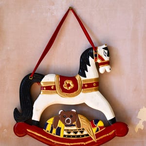 May include: A white rocking horse ornament with a black mane and tail, featuring red and gold saddle details. A small brown bear figure sits on the red rocker base, which is decorated with yellow, blue, and black patterns. A red ribbon is attached.