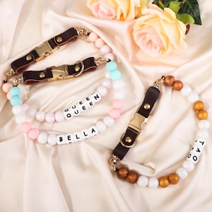 May include: Dog collars with brown leather straps and gold-coloured buckles. The collars are adorned with colourful beads in shades of pink, white, blue, and gold. One collar spells out "BELLA" and "QUEEN" with white letter beads.