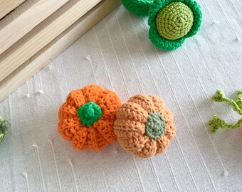 May include: Close-up of handmade crochet pumpkins in orange and tan with green stems. The pumpkins are on a white textured surface. Other crochet vegetables are also visible.